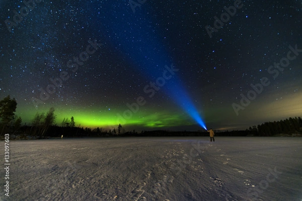 Fototapeta person puts bright blue flashlight to the starry sky with green north lights using torch staying on frozen lake 