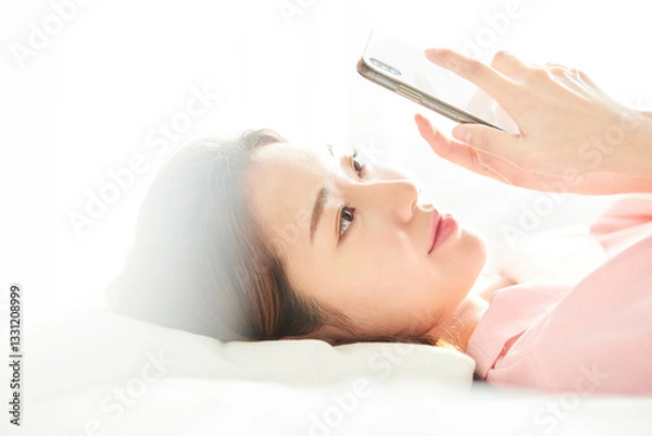 Fototapeta A young and beautiful woman, dressed in pink, is lying on a bed in a sunlit room, smiling happily while looking at her smartphone