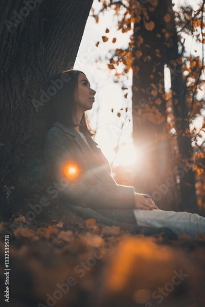 Fototapeta A young woman in a coat sits thoughtfully alone in an autumn forest against a background of fallen leaves. Autumn. Loneliness. Depression