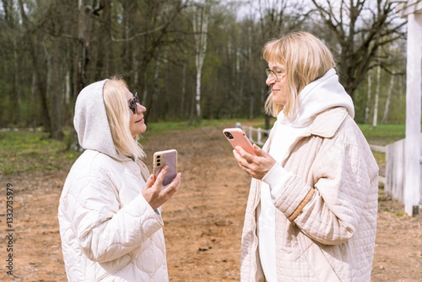 Fototapeta Two senior women standing in a park and talking while holding their smartphones. Dressed in stylish neutral-tone outfits with hoodies and sunglasses, they represent active aging and communication.
