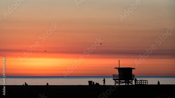 Fototapeta A beautiful sunset casts vibrant colors across a tranquil beach scene, highlighting silhouettes of people and iconic lifeguard station against the calming ocean backdrop.