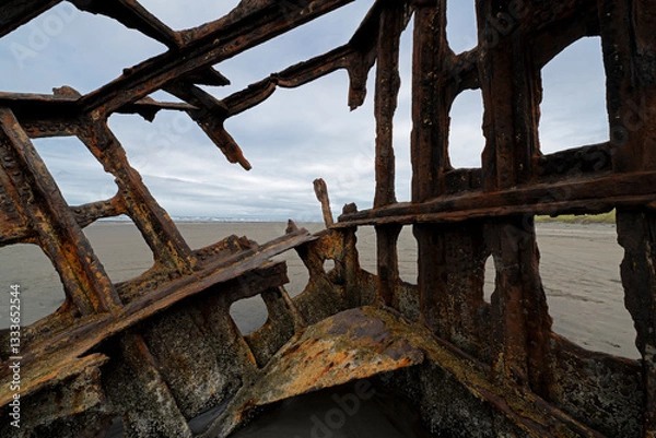 Fototapeta Inside the decaying hull of a shipwreck, the rusty structure tells stories of ocean adventures, surrounded by sandy shores and distant hills under an overcast sky.
