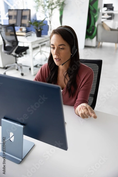 Obraz Serious online teacher giving business webinar on Internet. Busy Hispanic sales manager talking to company client on video chat, using wireless headset with mic and desktop monitor