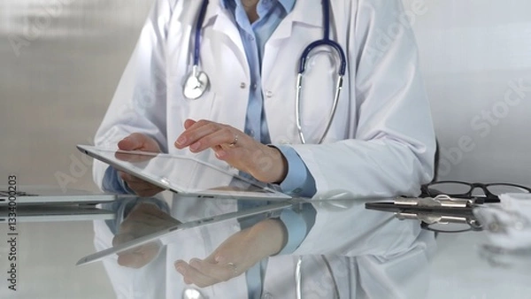 Fototapeta Doctor woman, wearing a blue blouse under a medical coat, is using digital tablet in modern healthcare setting, interacting with patient data, close-up. Medicine concept