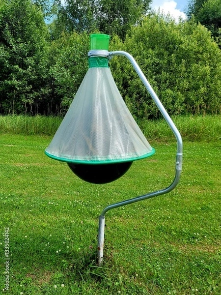 Fototapeta Horsefly trap against green trees. Photo of black ball insect trap on stand in mown meadow. Agricultural equipment for insect protection.