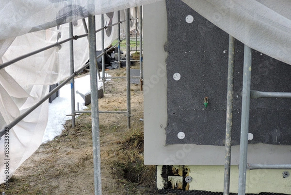 Obraz A wall of a house covered with EPS graphite polystyrene boards for thermal insulation, a scaffolding and building safety scaffolding net, some adhesive, anchors, an exterior wall mounted water faucet