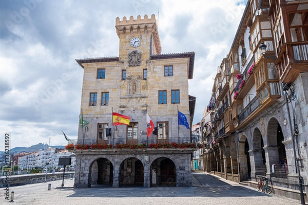 Obraz main facade of the town hall of Castrourdiales, with the flags waving in the wind, with its empty square of people, and its typical arcaded arcades, on a cloudy summer day