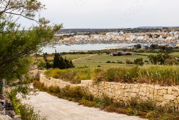Obraz Scenic countryside view of Marsaxlokk, Malta. A beautiful rural road in Malta leading towards Marsaxlokk Bay, featuring traditional stone walls, green fields, and a picturesque fishing village