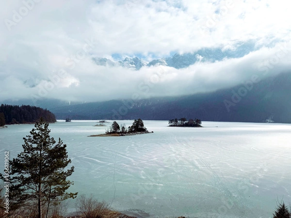 Fototapeta Scenic view over the frozen Eibsee lake with small islands with trees in the middle. The sky is covered by clouds but through a gap the Zugspitze mountain range is glimpsing through.