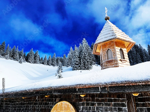 Fototapeta Low angle view of a wooden chapel in the mountains covered in snow against the blue sky with a mountain forest in the background
