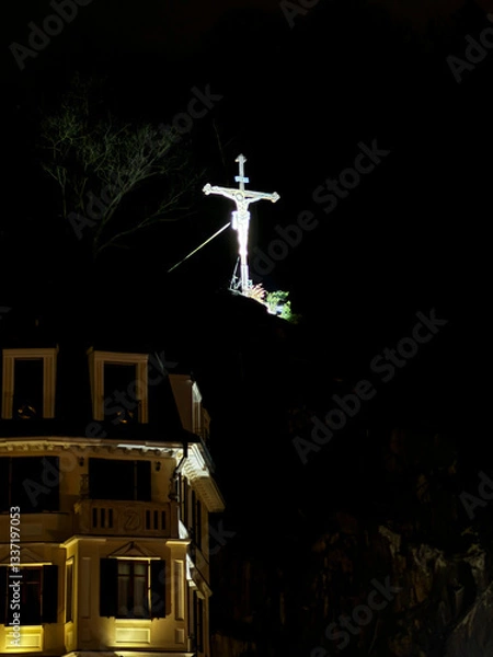 Fototapeta Old low-rise buildings of Karlovy Vary against the backdrop of a large illuminated cross at night
