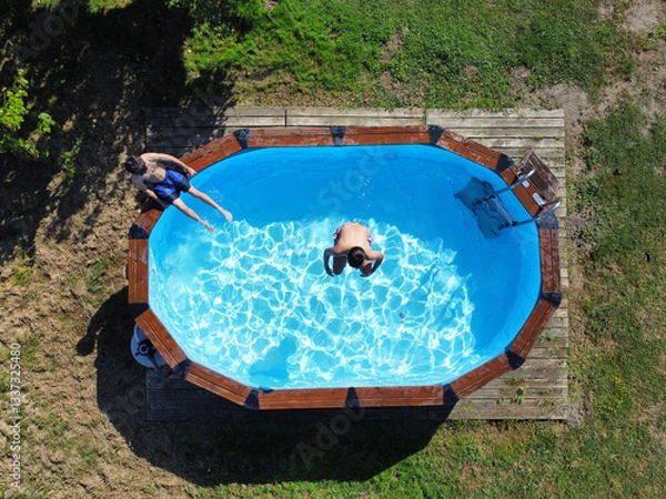 Obraz drone view of two teenagers enjoy a swim in a removable pool mounted on a wooden platform on the grass of a garden, drone view