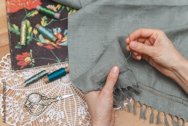Fototapeta Hands meticulously working on openwork embroidery on grey fabric, surrounded by vintage scissors, thread spools, and a floral book on a lace doily, showcasing a delicate crafting process.