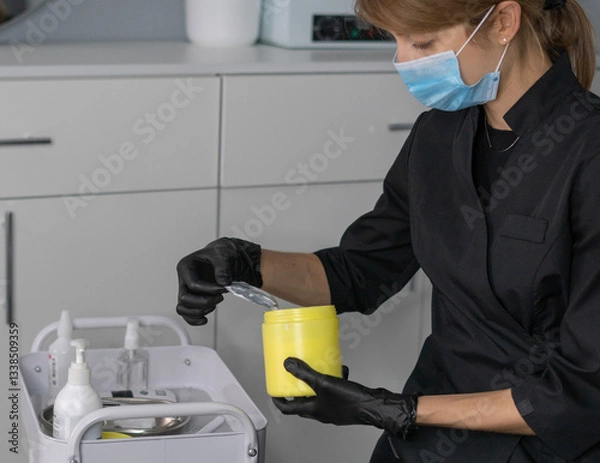 Fototapeta A cosmetologist with gloves and a mask carefully applies painkiller cream from a yellow container while preparing for a skincare treatment in a calm setting