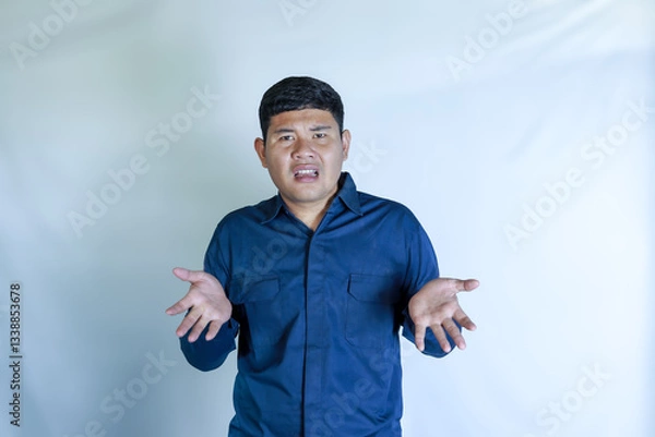 Obraz young asian man in blue shirt with confused expression with hand gesture, his face shows pain expression, headache with white studio background. Concept of disease, treatment, health care