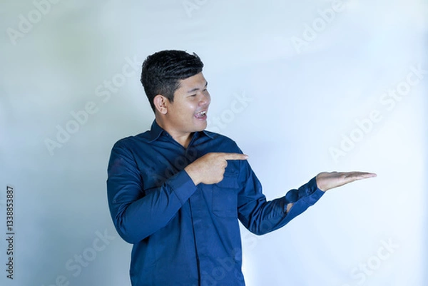 Obraz Handsome Asian Man Smiling in Blue Shirt Pointing to Empty Space in Hand with Cheerful Expression, Presenting Product, Idea or Advertising Message, Isolated on White Background