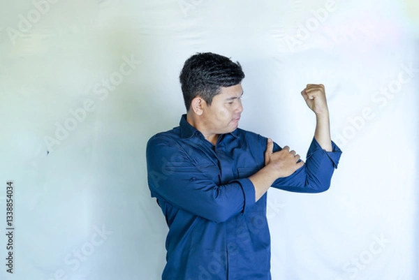 Obraz an asian man in blue shirt holding his hand on his bicep showing muscles on a white background