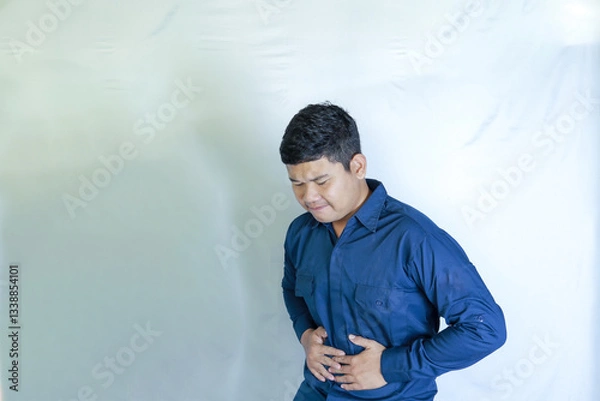 Obraz Young Asian man in blue shirt holding stomach with both hands, his face showing pain expression, stomachache with white studio background. Disease concept