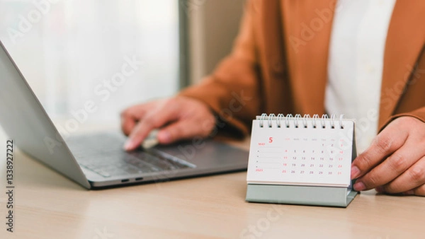 Fototapeta Monthly calendar May 2025 placed on office desk next to professional working with laptop, illustrating business schedule, daily planning, task management, deadlines, and productivity enhancement.