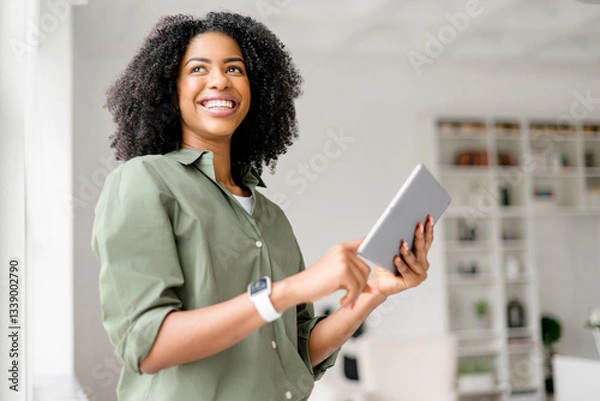 Fototapeta African-American woman is confidently handling a tablet, standing in a well-lit room with a smile. Cheerful female student or remote worker using digital tablet