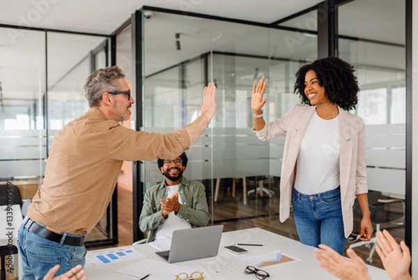 Fototapeta A team leader and a businesswoman exchange a high-five in a collaborative office setting while teammates cheer. A representation of teamwork, celebration, and motivation in the corporate world.