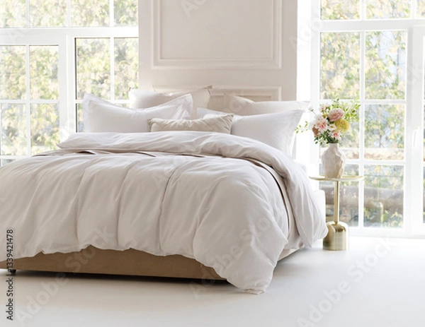 Fototapeta Sunny bedroom with two large windows. Four pillows, cushion and a fluffy down comforter on a double bed with bedside table and bouquet in vase on it. Side view horizontal photo. Suitable for mockup