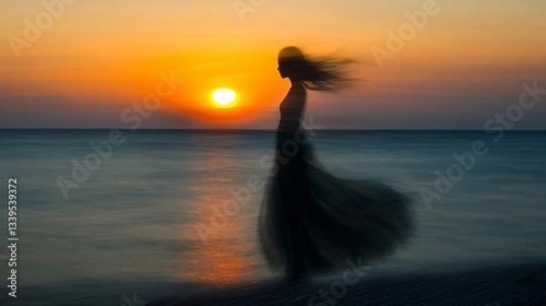 Fototapeta A blurred silhouette of a woman in a dress stands on a beach at sunset, with the sun setting over the ocean.