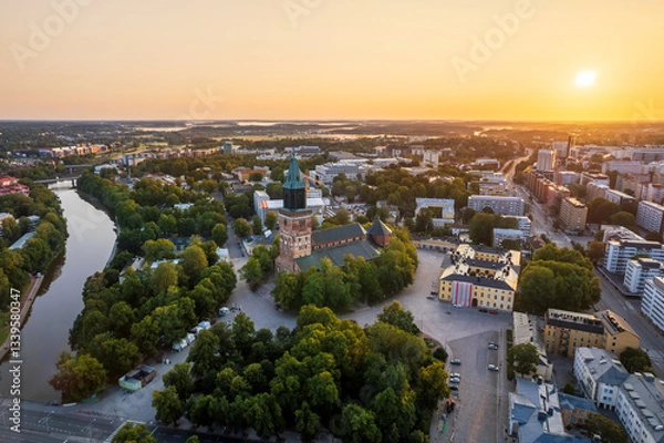 Fototapeta Aerial view of Turku Cathedral and the surrounding cityscape at sunrise. The historic church, built in the late 13th century, stands by the Aura River, surrounded by lush greenery and urban landmarks.