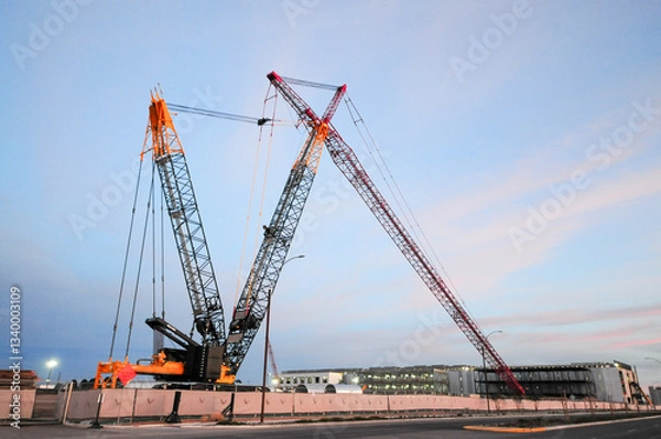 Fototapeta Large crawler cranes with lattice booms positioned at a data center construction site, supporting heavy lifting operations for structural assembly, with buildings under construction in the background
