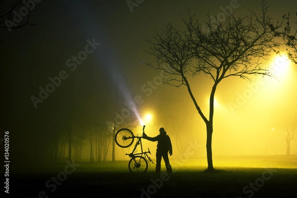 Fototapeta One man is standing and holding the bicycle in foggy and mysterious park at night