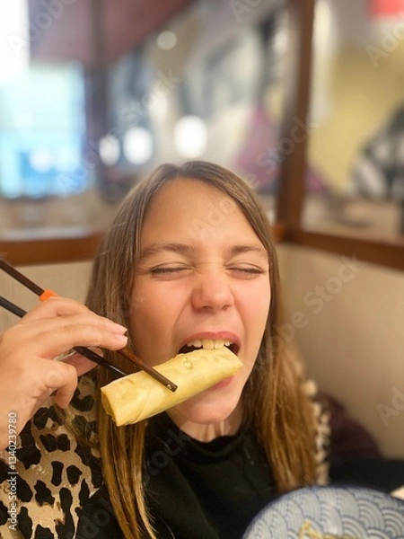 Obraz A young girl bites into a crispy spring roll using chopsticks at an Asian restaurant, enjoying her meal with an expressive face.