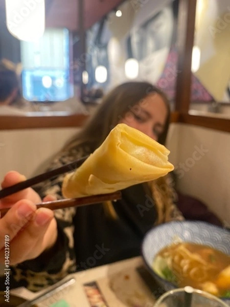 Obraz A young girl in a restaurant holds a freshly made spring roll with chopsticks, offering a close-up view of the food.