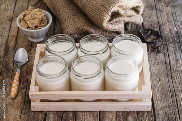 Obraz Homemade yogurt in glass jar and wooden box. Antique wooden table. Burlap and cereal bowl on background. Steel teaspoon orange and white dots