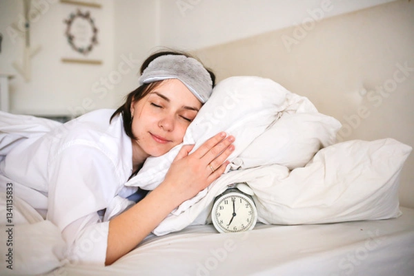 Fototapeta Young woman in a sleep mask holding an alarm clock while lying on a bed in a bright room, symbolizing a healthy wake-up routine.