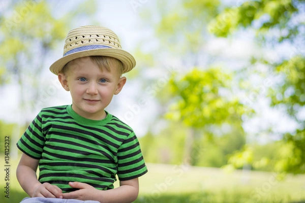 Fototapeta Young boy sitting in the grass at the park wearing a fedora