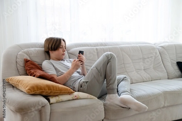 Fototapeta Teenager works on homework while relaxing on a couch in a cozy living room during the afternoon. Full length portrait of young teenage boy in hand smartphone while sitting on sofa