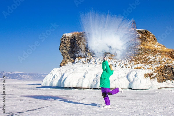 Fototapeta Girl in colorful clothes splashes hot water in air at cold winter day while on frozen Baikal lake. The droplets immediately freeze, turn into steam and ice crystals. Mpemba effect or paradox. Russia.