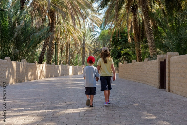 Fototapeta Two kids walk through Al Ain Oasis, UAE, a popular tourist spot. The oasis offers shade and a cool escape from the heat, making it a perfect place for a relaxing walk in nature