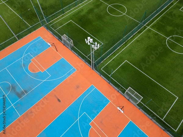 Fototapeta Aerial view of outdoor basketball and soccer courts with vibrant blue, orange, and green colors. The sports facility is empty, with clear markings and fences, creating a dynamic geometric composition