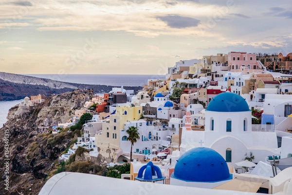 Fototapeta Scenic view of Oia, Santorini, Greece, with blue-domed churches, colorful houses, and the Aegean Sea. Horizontal shot with a cloudy sky, a beautiful island travel, honeymoon or wedding destination