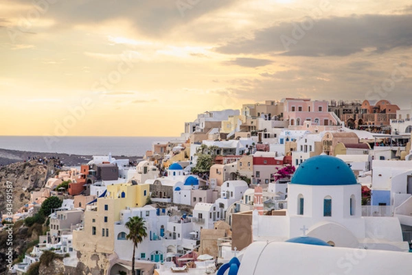 Fototapeta Scenic view of Oia, Santorini, Greece, with blue-domed churches, colorful houses, and the Aegean Sea. Horizontal shot with a cloudy sky, a beautiful island honeymoon or wedding destination