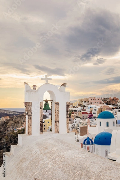 Fototapeta Scenic view of Oia, Santorini, Greece, featuring the iconic three domes viewpoint, colorful houses, and the Aegean Sea. Vertical shot with a cloudy sky, perfect for honeymoon or wedding destination