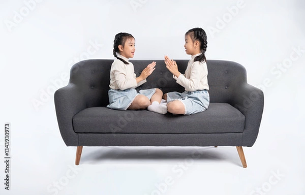 Fototapeta Two young girls sitting face-to-face on a gray couch, playing a clapping game, against a white backdrop.