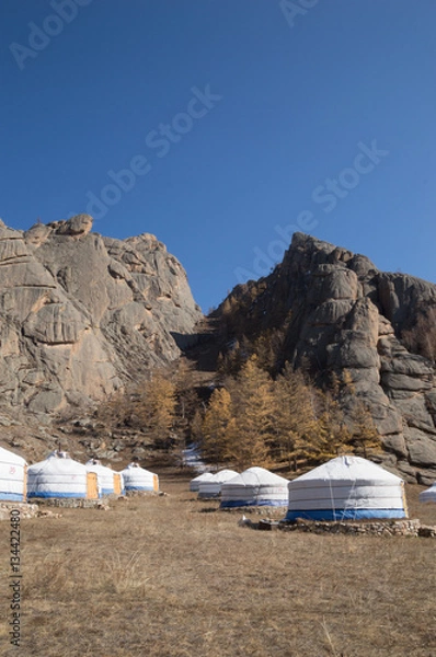 Fototapeta White and blue canvas gers at the base of rugged mountains. The yurts are on stone foundations. Sparse vegetation is in the background. Deep blue sky.