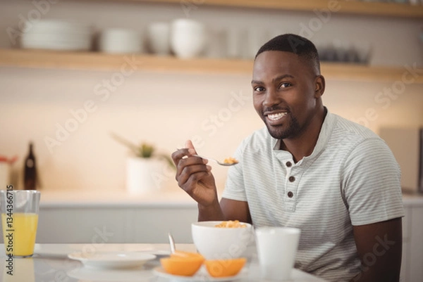 Fototapeta Smiling man enjoying breakfast cereal at home with orange juice on table, copy space