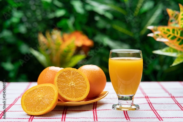 Obraz Glass cup filled with orange juice next to a yellow plate with cut oranges against a background of green plants