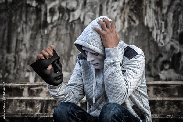Fototapeta Asian man in the hood and white mask with confused depression sitting on stairs and looking at black mask, in scary abandoned building, Human face expression, Good and Bad temptation concept.