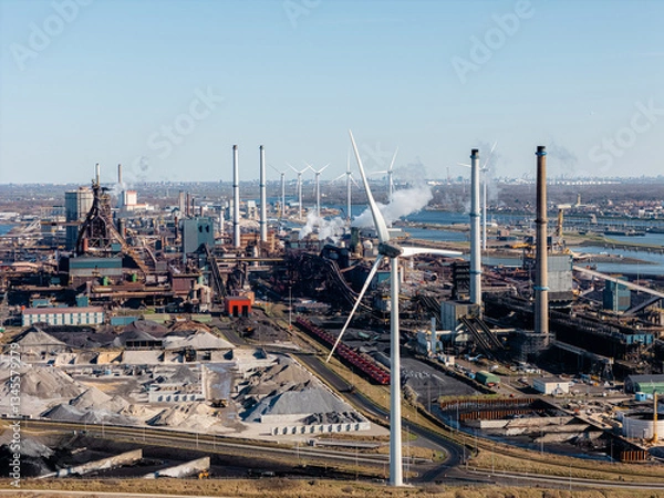 Obraz Steel production site in IJmuiden, Netherlands, featuring smokestacks, raw materials, and wind turbines. The aerial perspective highlights the tension between heavy industry and renewable energy.