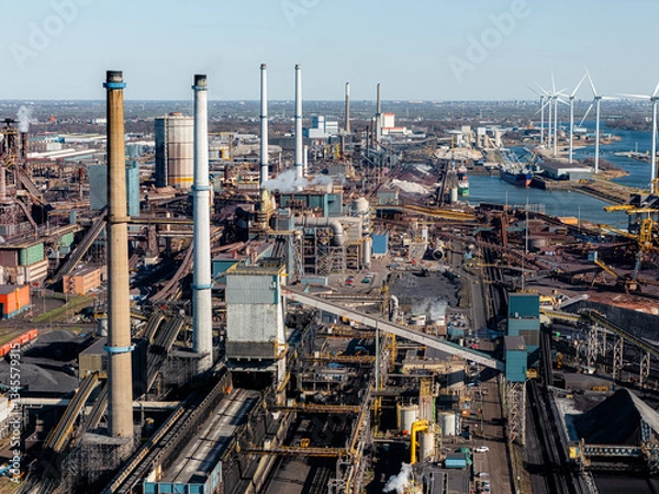 Obraz Industrial steel plant in IJmuiden, Netherlands, with large chimneys, conveyor belts, and emissions. Aerial view of a major production hub, showing both traditional and renewable energy elements.
