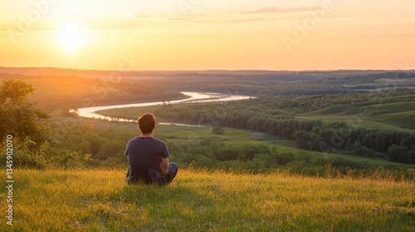 Fototapeta Serene sunset view, man meditates atop grassy hill overlooking winding river valley